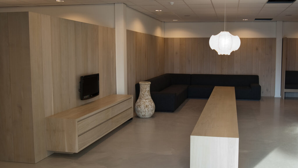 Moderne wachtruimte met houten wand, zwarte hoekbank en lange houten tafel bij Tandarts Alkmaar Oudorp