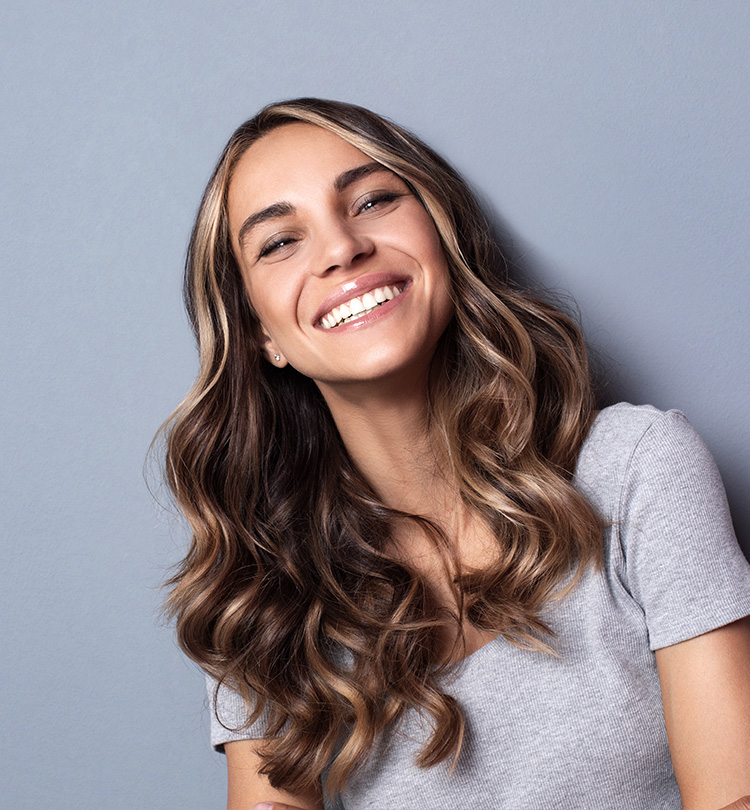 Vrouw met lang bruin haar die breed glimlacht voor een grijze achtergrond